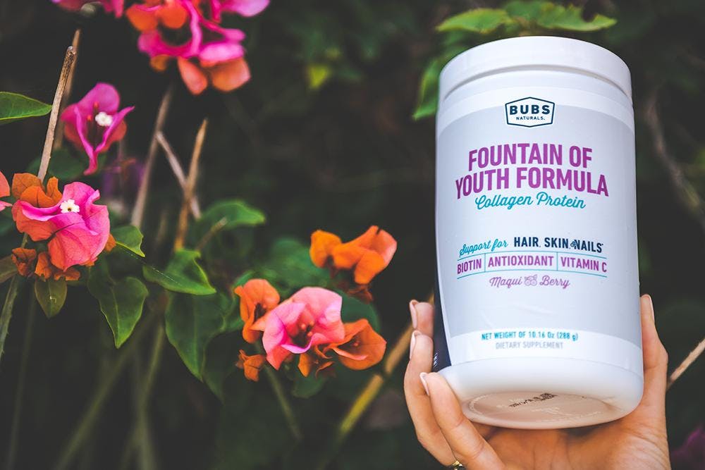 a woman holding BUBS Naturals Fountain of Youth Formula Collagen Protein in front of flowers outside