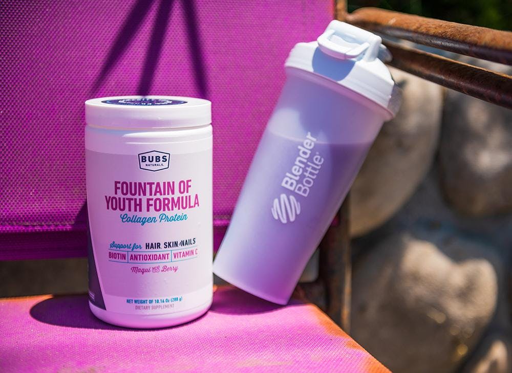 BUBS Naturals Fountain of Youth Formula Collagen Protein container next to a Blender Bottle filled with a protein shake on a chair