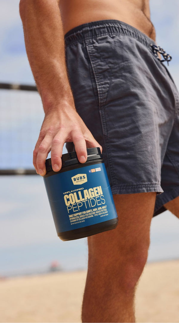 A man wearing dark gray swim trunks holds a blue tub of Bubs Collagen Peptides on a sandy beach volleyball court.